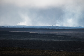 A lava plain with dark volcanic rock and low ridges extends in Iceland. Steam rises from fissures on the Reykjanes Peninsula in soft overcast light, evoking raw geology.