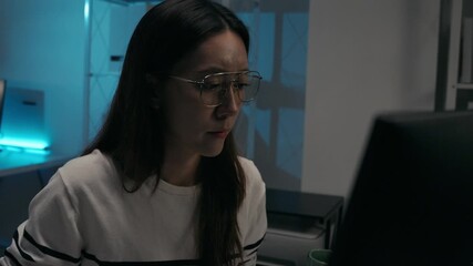 Asian woman IT professional adjusting her glasses while working late on a computer. Deep concentration or eye strain from long hours of remote work or coding in a dark office