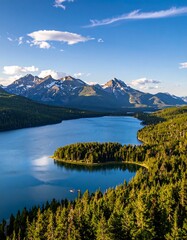 Aerial view of a pristine lake surrounded by evergreen forest, with majestic, snow-capped mountains under a bright, blue sky