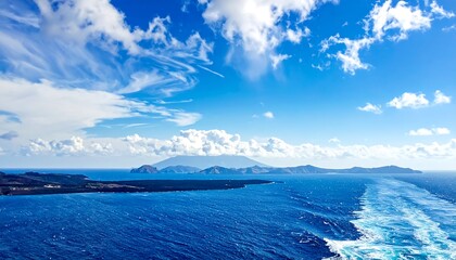 Aerial view of a vast, clear blue sea under a bright sky with puffy clouds, and a distant island range on the horizon. A wake trails from the ship