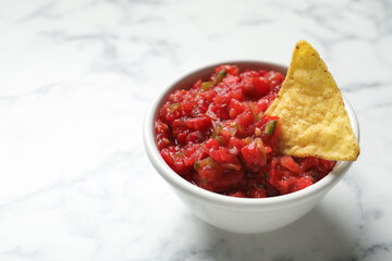 Tasty salsa in bowl and nacho on white marble table, closeup. Space for text