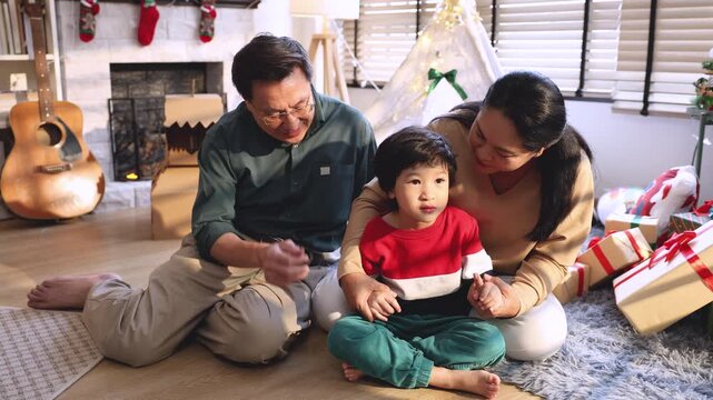 Asian grandfather, grandmother, and cheerful grandson sitting on floor in cozy living room, exchanging smiles and enjoying warm bonding moments together during Christmas gift time, festival.