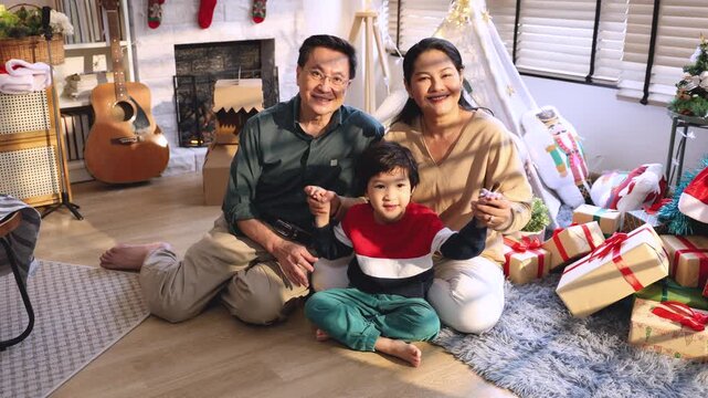 Asian grandfather, grandmother, and cheerful grandson sitting on floor in cozy living room, exchanging smiles and enjoying warm bonding moments together during Christmas gift time, festival.