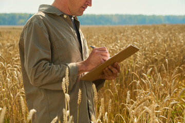 Young man standing in wheat field writing on clipboard, conducting agricultural inspection or research, focusing on crop assessment during harvest season