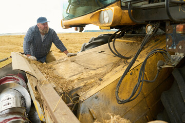 Middle aged Caucasian man inspecting large agricultural machine in wheat field during harvest season, kneeling beside equipment and examining machinery components closely © pressmaster