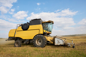 Obraz premium Yellow combine harvester operating in wheat field during harvest season, large agricultural machine moving through golden crop under blue sky with scattered clouds, rural landscape visible