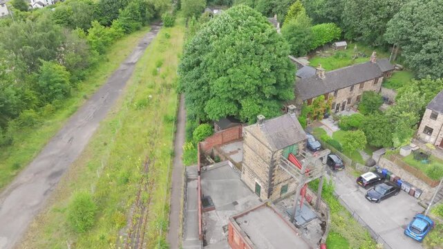 Ascending drone orbit over Elsecar Heritage Centre showing stone and brick workshops, sawtooth roofs, the New Yard chimney, central market tents, parked vehicles and an overgrown disused railway.