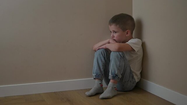 Sad child sits on floor in corner arms folded across chest. Child posture remains still as sadness fills quiet space. Emotion reflects child face. Corner creates soft frame around deep inner sadness.