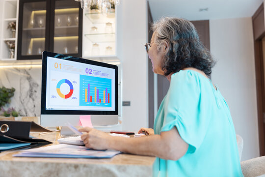 An elderly woman works remotely from home, analyzing colorful charts and graphs on her computer screen. Concept of senior digital skills, remote data analysis, and active aging through technology.