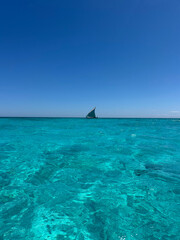 Obraz premium A vast, sparkling turquoise ocean stretches toward the horizon in Madagascar, with a small sailboat visible in the distance. The bright sunlight reflects off the calm water, creating a serene and idyl