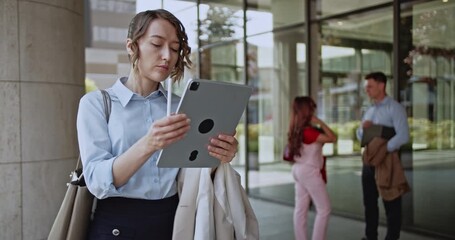 A businesswoman stands outside a sleek office building, diligently working on her digital tablet. Her professional demeanor reflects modern corporate efficiency and connectivity in an urban setting. - Powered by Adobe