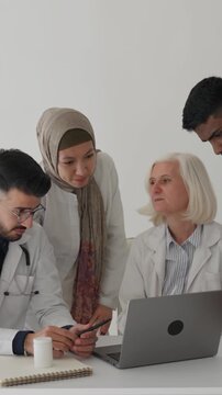 Diverse team of physicians and medical students in white coats gathered around a laptop in a hospital, collaborating, discussing a patient case and analyzing diagnostics together