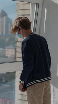 Young male patient wearing a protective face mask looking at window and waving parents in hospital room, feeling lonely and isolated while looking at the city outside during quarantine