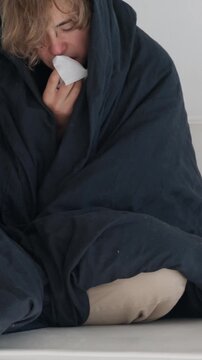Sick young man with blonde hair wrapped in a dark blanket feeling unwell, sneezing, and wiping his running nose with a tissue, isolated on a white background. Seasonal illness and health concept