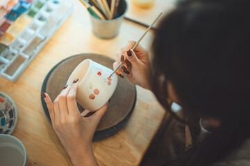 Close-up of female artist’s hands sketching design with pencil on handmade ceramic pottery before painting – creative hobby, traditional craft and artistic expression in calm studio workshop setting