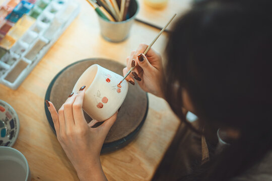Close-up of female artist’s hands sketching design with pencil on handmade ceramic pottery before painting – creative hobby, traditional craft and artistic expression in calm studio workshop setting