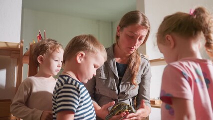 Coach introduces children to tortoise as they gather curiously, one child wearing hearing aid in striped shirt watches closely, while others observe interaction with focus in classroom setting
