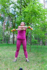 A fit mature woman holding dumbbells while performing an exercise in a green park, staying active and healthy.