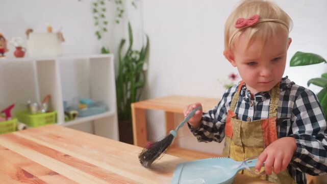 Beautiful kid wearing checkered shirt and apron using brush to sweep flour into parker on wooden table inside bright kindergarten environment with shelves, plants, and kitchen utensils in background