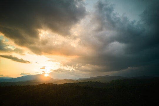 Aerial view of dramatic sky with thick, dark clouds looms over mountainous landscape. Sun peeks through, casting warm, golden light on rolling hills and dense forests below, creating striking contrast
