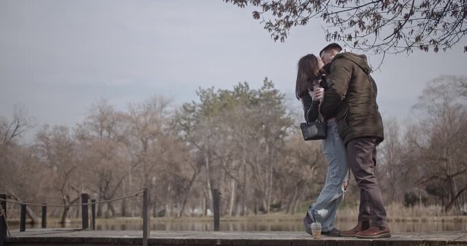 A loving young couple shares tender kisses and warm hugs by the tranquil lake in a beautiful park. Their intimate moments showcase deep affection and companionship on a peaceful day.