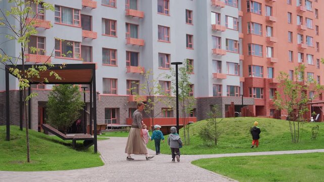 Teacher walks with group of young children through green residential courtyard surrounded by colourful apartment buildings, guiding them safely during outdoor activity under cloudy sky