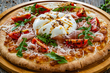 Pizza with cured ham, burrata cheese, tomatoes and arugula on wooden table