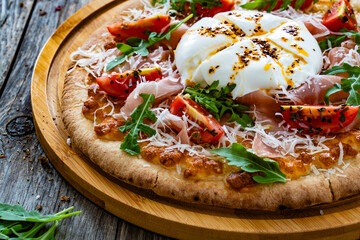 Pizza with cured ham, burrata cheese, tomatoes and arugula on wooden table