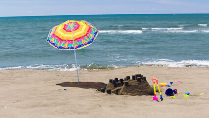 Sandcastle and colorful beach toys beside a bright parasol on the sunny shore, waves lapping the...