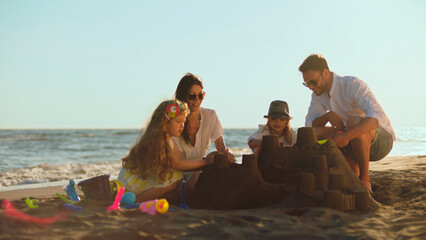 Happy family of four enjoying summer beach vacation, parents with two young children building a sandcastle on the sand by the ocean at sunset, fostering togetherness and fun