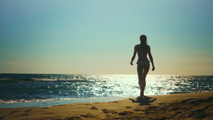 Woman in bikini walking along a sandy shoreline at golden hour, silhouetted against the sparkling sea and warm sunset glow, enjoying peaceful solitude and freedom