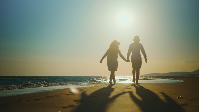 Two children silhouetted holding hands, walking barefoot along a sandy beach at sunset, long shadows stretching across the shore as waves lap the tranquil ocean horizon - Powered by Adobe