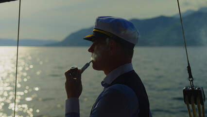 Sailor man wearing a captain's hat and vest, standing on a sailboat, smoking a pipe and enjoying a...