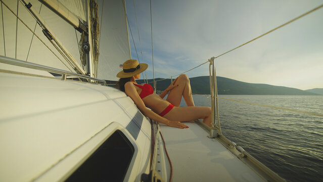 Woman in a red bikini and straw hat relaxing on the deck of a sailing boat, enjoying the summer sun and tranquil ocean views during a leisurely vacation - Powered by Adobe