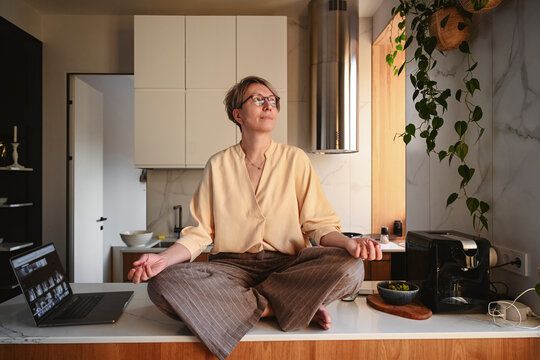 Woman in glasses meditating on a kitchen table with a laptop nearby