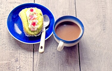 Dessert: The ingredients consist of a bright lime green roll cake topped with white frosting, sprinkles of sugar and a shiny red cherry. The cake is placed on a blue plate with a coffee cup next to it