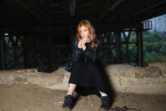 Person in leather jacket and red sweater sitting under a bridge outdoors