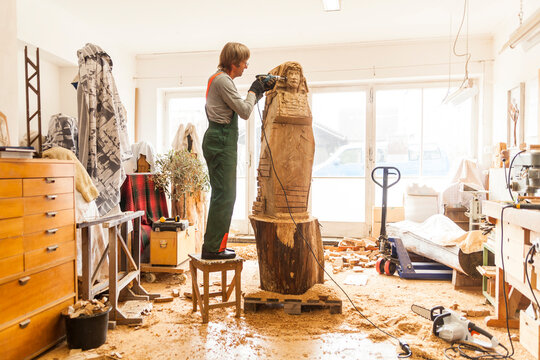 Wood carver in workshop working on sculpture with milling machine, standing on stool