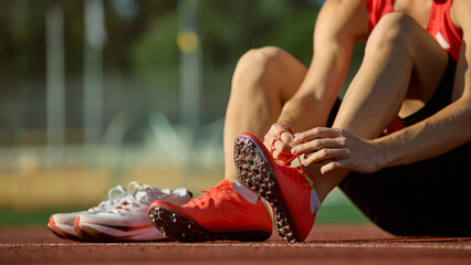 Close-up of runner tying red spikes on running track under sunlight. Concept of preparation, detail, discipline for sports branding, footwear advertising, and athletic motivation visuals.