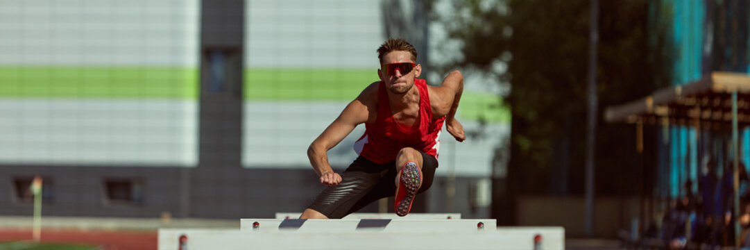 Runner in red outfit clearing hurdle during sprint on athletic field. Concept of effort, discipline, mental strength for motivation, fitness branding, and sport promotion. - Powered by Adobe