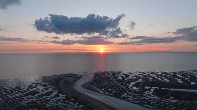 Setting Sun On Horizon Over Calm Irish Sea With Reverse Camera Flight Over Dark Tidal Sand Flats And Inlets. Golden Hour. Fleetwood, Lancashire, UK.