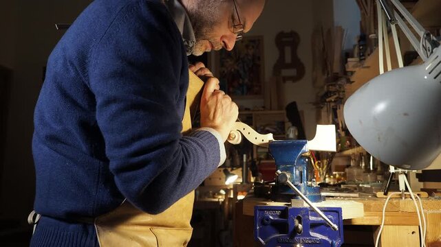 Master luthier leans over his workbench carving the scroll of a violin by hand under bright lamplight, the focused posture and warm tones capture the dedication artistry of traditional violin making.