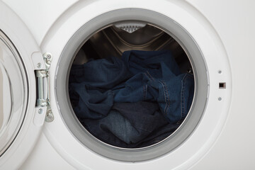 Dark blue denim jeans in opened white washing machine. Clothes care. Closeup. Front view.