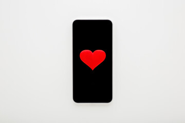 Smartphone with dark black touch screen and red heart shape on light gray table background. Love concept. Online dating. Closeup. Top down view.