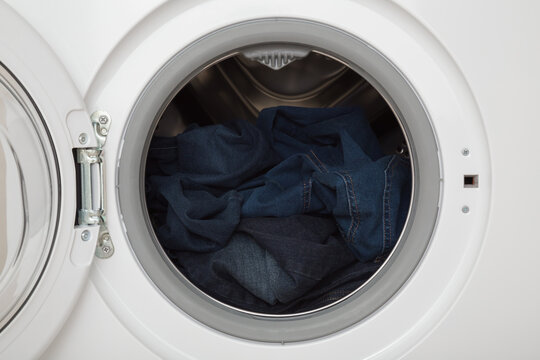 Dark blue denim jeans in opened white washing machine. Clothes care. Closeup. Front view.
