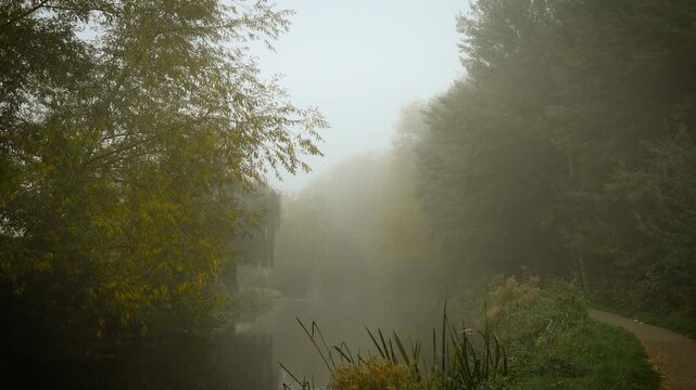 Misty morning covering calm river with narrow path along shore. Creeping mist wrapping ancient path forming mysterious feeling. Foggy dawn veiling still water beside trail bordered by autumn woods