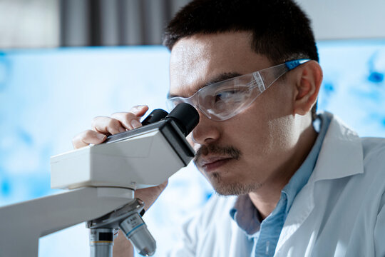 A man wearing a lab coat and safety glasses is looking through a microscope