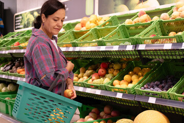 Cheerful 40-year-old woman in casual clothes shops for daily groceries, food items, and other items along the aisles and shelves of grocery store. Healthy eating.