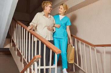 Compassionate nurse caregiver helps elderly senior woman descend home stairs, offering patient support and assistance for confidence and family contact. Nurse helps elderly woman walk down stairs.