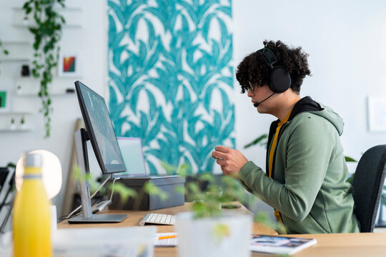 Side view of Arab young male in hoodie and headset holding gamepad while playing videogame on computer at desk in cozy room